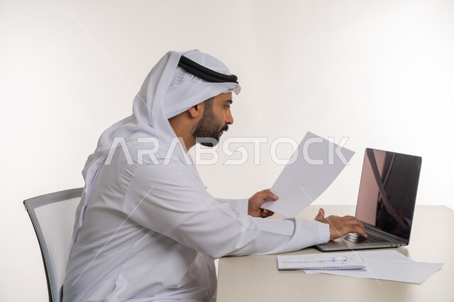 العمل عن بعد باستخدام جهاز الحاسوب المحمول في الأعمال المكتبيه, بورتريه مقرب من الجانب لرجل عربي خليجي اماراتي يرتدي الغترة والكندورة البيضاء ينظر الى جهاز اللابتوب بايماءات التركيز, مهن ووظائف مكتبية, خلفيه بيضاء