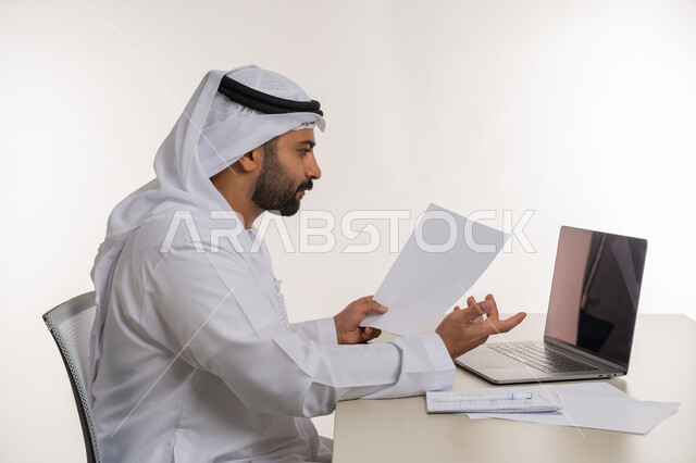 Remote work using laptop in office work, portrait of an Arab Gulf Emirati man wearing a white ghutra and kandura raising his hand in a gesture of surprise and dissatisfaction, office jobs and professions, white background