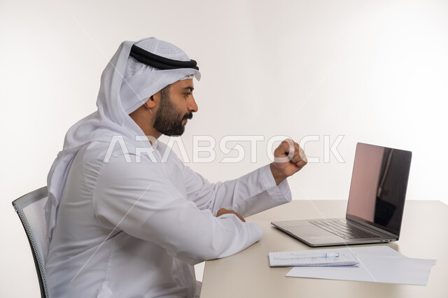العمل عن بعد باستخدام جهاز الحاسوب المحمول في الأعمال المكتبيه, بورتريه مقرب من الجانب لرجل عربي خليجي اماراتي يرتدي الغترة والكندورة البيضاء يرفع قبضة يده بايماءات الفرح بالنجاح والفوز, مهن ووظائف مكتبية, خلفيه بيضاء