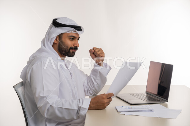 العمل عن بعد باستخدام جهاز الحاسوب المحمول في الأعمال المكتبيه, بورتريه مقرب من الجانب لرجل عربي خليجي اماراتي يرتدي الغترة والكندورة البيضاء ينظر الى الورقة بايماءات الفرح بالنجاح والفوز, مهن ووظائف مكتبية, خلفيه بيضاء