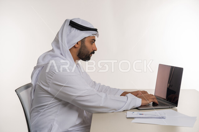 العمل عن بعد باستخدام جهاز الحاسوب المحمول في الأعمال المكتبيه, بورتريه مقرب من الجانب لرجل عربي خليجي اماراتي يرتدي الغترة والكندورة البيضاء ينظر الى جهاز اللابتوب بايماءات التركيز, مهن ووظائف مكتبية, خلفيه بيضاء