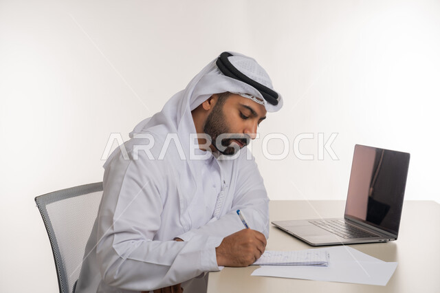 انجاز المهام وتحقيق الاهداف, بورتريه مقرب من الجانب لرجل عربي خليجي اماراتي يرتدي الغترة والكندورة البيضاء يقوم  بتدوين الملاحظات ومراجعتها بايماءات التركيز, استخدام جهاز الحاسوب المحمول, خلفية بيضاء