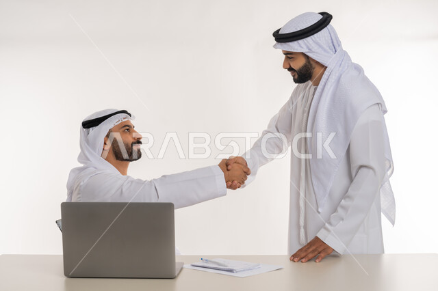 مهن ووظائف مكتبية, مفهوم التعاون بين الموظفين, التفاعل الجماعي لتحقيق أهداف الشركة, دمج التقنيات الحديثة المتطورة في الحياة العملية, بورتريه لموظفين عربيين خليجيين اماراتيين مبتسمين يرتديان الغترة والكندورة البيضاء يتصافحان باليدين, خلفية بيضاء