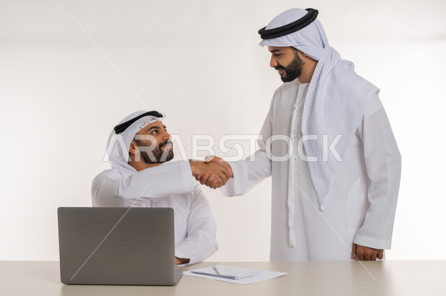 مهن ووظائف مكتبية, مفهوم التعاون بين الموظفين, التفاعل الجماعي لتحقيق أهداف الشركة, دمج التقنيات الحديثة المتطورة في الحياة العملية, بورتريه لموظفين عربيين خليجيين اماراتيين مبتسمين يرتديان الغترة والكندورة البيضاء يتصافحان باليدين, خلفية بيضاء