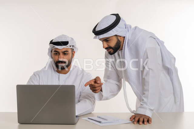 مفهوم التعاون بين الموظفين, التفاعل الجماعي لتحقيق أهداف الشركة وحل المشكلات, دمج التقنيات الحديثة المتطورة في الحياة العملية, رجل أعمال عربي خليجي اماراتي يرتدي الغترة والكندورة البيضاء يقف بجانب زميله يشير باصبع السبابة الى جهاز الحاسوب المحمول, خلفية بيضاء