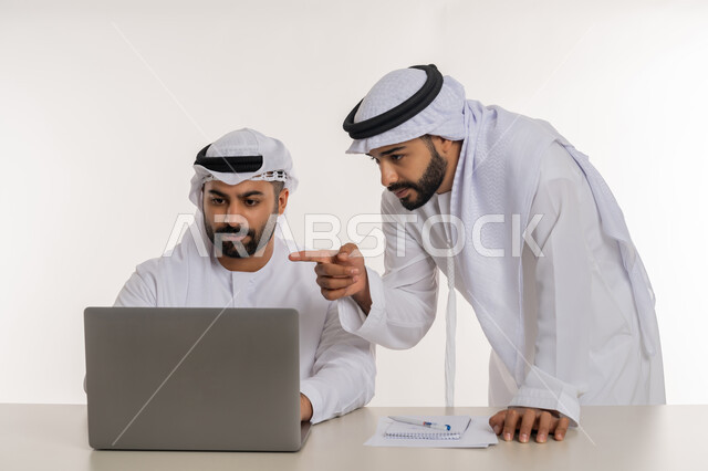 مفهوم التعاون بين الموظفين, التفاعل الجماعي لتحقيق أهداف الشركة وحل المشكلات, دمج التقنيات الحديثة المتطورة في الحياة العملية, رجل أعمال عربي خليجي اماراتي يرتدي الغترة والكندورة البيضاء يقف بجانب زميله يشير باصبع السبابة الى جهاز الحاسوب المحمول, خلفية بيضاء
