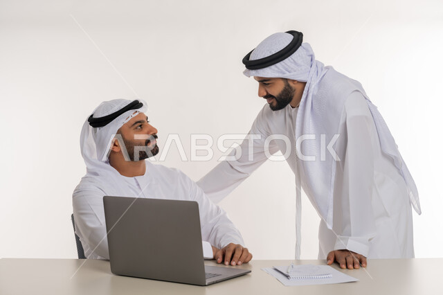 Unity and team interaction to achieve company goals and solve problems, portrait of two Emirati Gulf Arab employees wearing white ghutra and kandura discussing work matters, office jobs and professions, white background
