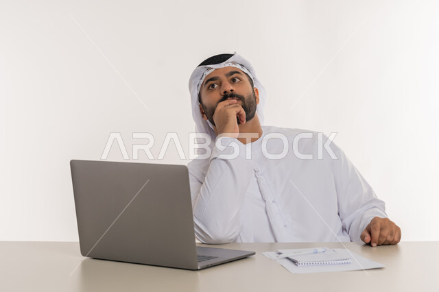 تعابير التفكير في استراتيجيات وأهداف العمل، بورتريه مقرب لرجل عربي خليجي اماراتي يرتدي الغترة والكندورة البيضاء  يضع يده على ذقنه ينظر الى شيء ما بايماءات شرود الذهن والحيرة، تطور التكنولوجيا وإستخدام الأجهزة التقنية الحديثة، مهن ووظائف مكتبية, خلفية بيضاء