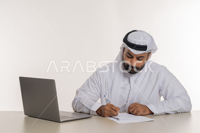 انجاز المهام وتحقيق الاهداف, بورتريه مقرب لرجل عربي خليجي اماراتي يرتدي الغترة والكندورة البيضاء يقوم  بتدوين الملاحظات ومراجعتها بايماءات التركيز, استخدام جهاز الحاسوب المحمول, خلفية بيضاء
