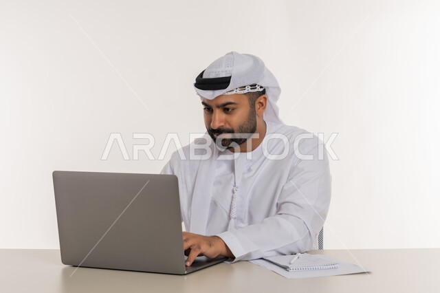العمل عن بعد باستخدام الحاسوب المحمول في الأعمال المكتبيه, بورتريه مقرب لشاب عربي خليجي اماراتي يرتدي الغترة والكندورة البيضاء  ينظر الى جهاز اللابتوب بايماءات التركيز, خلفيه بيضاء