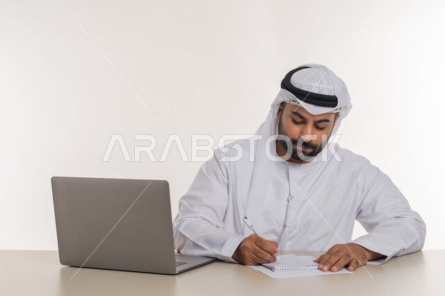 انجاز المهام وتحقيق الاهداف, بورتريه مقرب لرجل عربي خليجي اماراتي يرتدي الغترة والكندورة البيضاء يقوم  بتدوين الملاحظات ومراجعتها بايماءات التركيز, استخدام جهاز الحاسوب المحمول, خلفية بيضاء