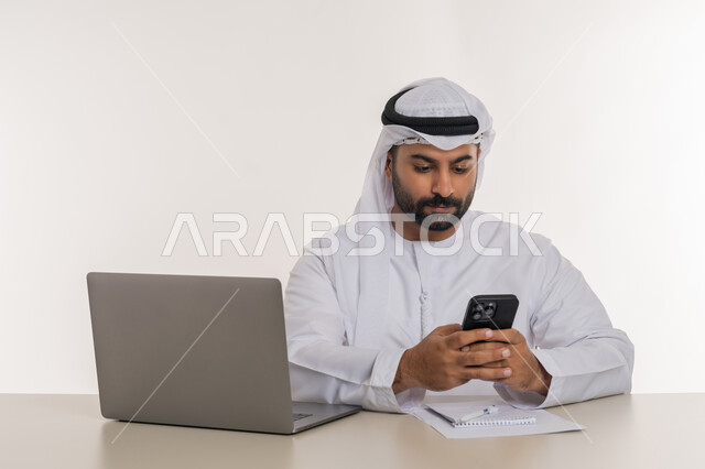 مهن ووظائف مكتبية، بورتريه مقرب لرجل عربي خليجي اماراتي يرتدي الغترة والكندورة البيضاء يحمل بيده الجوال وينظر اليه بايماءات التركيز، تطور التكنولوجيا وإستخدام جهاز الحاسوب المحمول لإنجاز المهام, خلفية بيضاء