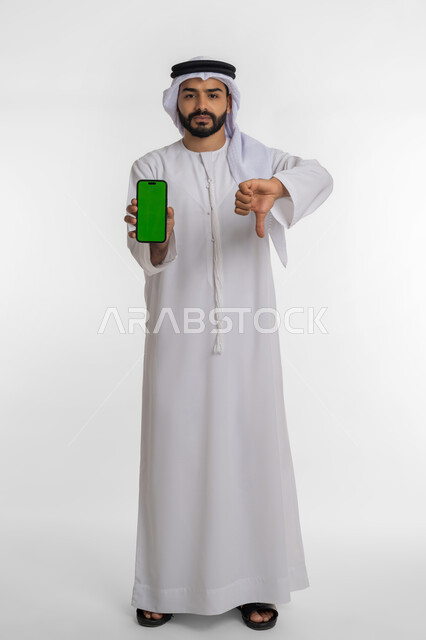 مفهوم التقييم السلبي والفشل في تحقيق أمر ما، استخدام التقنيات المتطورة,  بورتريه لرجل عربي خليجي إماراتي يرتدي الكندورة والغترة يمسك بيده الهاتف المحمول بشاشة خضراء فارغة يشير بإصبع الابهام الى الاسفل بإيماءات الرفض، صورة الجسم بالطول الكامل، خلفية بيضاء