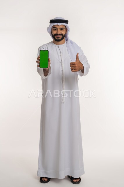 تعابير الإعجاب والرضا، استخدام التكنولوجيا والتقنيات الحديثة والمتطورة، بورتريه لرجل عربي خليجي إماراتي يرتدي الغترة و الكندورة يرفع اصبع الابهام للاعلى بإيماءات القبول والموافقة ويحمل الهاتف المحمول بشاشة خضراء فارغة، الجسم بالطول الكامل, خلفية بيضاء