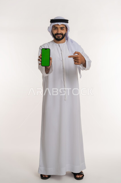 استخدام التكنولوجيا والتقنيات الحديثة والمتطورة، النظر الى الكاميرا بتعابير الإعجاب والرضا، بورتريه لرجل عربي خليجي إماراتي يرتدي الغترة و الكندورة يشير باصبع السبابة الى  الهاتف المحمول بشاشة خضراء فارغة، الجسم بالطول الكامل, خلفية بيضاء