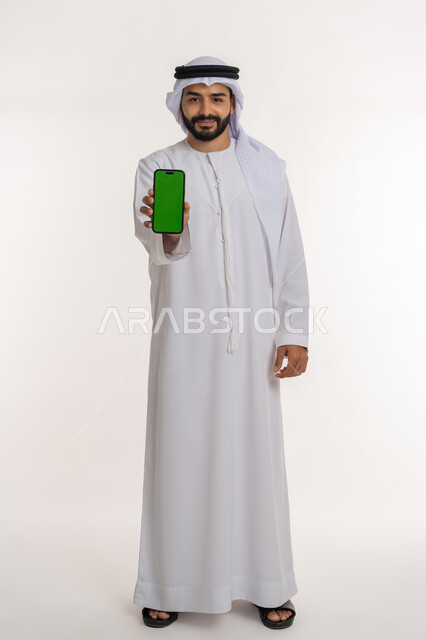 Looking at the camera with expressions of admiration and satisfaction, using modern and advanced technology and techniques, portrait of an Arab Gulf Emirati man wearing a ghutra and kandura holding a mobile phone with a blank green screen, full-length body, white background