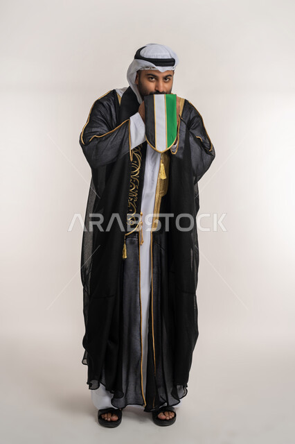 الاحتفال باليوم الوطني 2 ديسمبر، مفهوم حب الوطن والانتماء إليه، إحياء ذكرى استقلال البلاد، بورتريه لرجل عربي خليجي إماراتي يرتدي البشت يقوم بتقبيل علم الإمارات المتحدة، إيماءات الاعتزاز والفخر، صورة الجسم بالطول الكامل، خلفية بيضاء