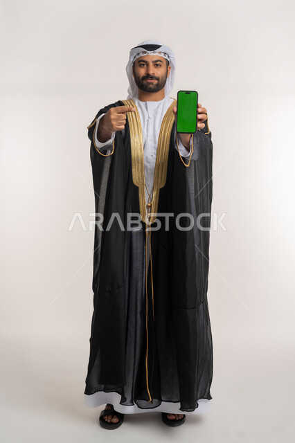 التسويق الإلكتروني وعرض خدمات وتطبيقات الجوال، بورتريه لرجل عربي خليجي إماراتي يرتدي البشت ويشير بإصبع السبابة نحو الهاتف المحمول بشاشة خضراء فارغة، إستخدام برامج الهاتف الذكي، الجسم بالطول الكامل, خلفية بيضاء