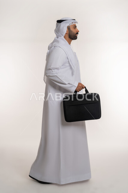 The concept of elegance and interest in external appearance, standing straight with expressions of self-confidence, portrait of an Arab Gulf Emirati man wearing a white kandura and ghutra, carrying a bag in his hand and standing with masculine gestures, full-length body, white background