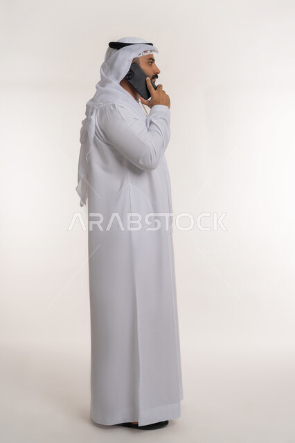 استخدام التقنيات والأجهزة الحديثة، التواصل مع الأهل والأصدقاء، بورتريه لرجل عربي خليجي إماراتي مبتسم يرتدي الكندورة والغترة يقوم بإجراء مكالمة هاتفية عبر الهاتف المحمول، الطول الكامل للجسم، خلفية بيضاء