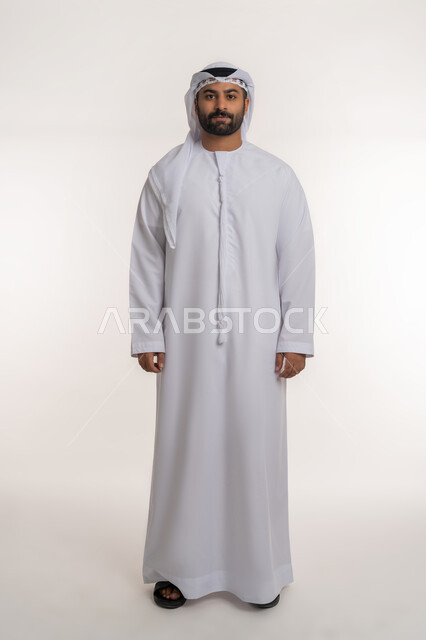 The concept of elegance and interest in external appearance, standing straight and looking at the camera with expressions of self-confidence, portrait of an Arab Gulf Emirati man wearing a white kandura and ghutra standing with gestures of manhood, full-length body, white background