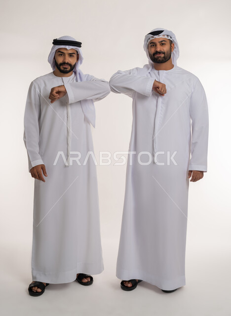 الحفاظ على المسافات بين الاشخاص, التباعد واتباع احترازات السلامة للوقاية من فايروس كورونا، بورتريه لرجلين عربيين خليجيين اماراتيين يرتديان الغترة والكندورة يقومان بالتصافح بقبضة اليد، الجسم بالطول الكامل, خلفية بيضاء