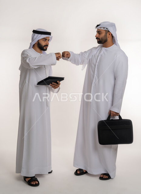 التباعد واتباع احترازات السلامة للوقاية من فايروس كورونا، الحفاظ على المسافات بين الاشخاص, بورتريه لرجلين عربيين خليجيين اماراتيين يرتديان الغترة والكندورة يقومان بالتصافح بقبضة اليد، الجسم بالطول الكامل, خلفية بيضاء
