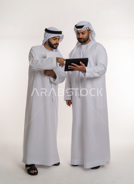 Using advanced modern equipment, the concept of collaborative management, a portrait of two Emirati Gulf Arab employees wearing ghutra, kandura and masks discussing using papers, achieving company goals, solving problems and making new partnership deals, full-length body, white background