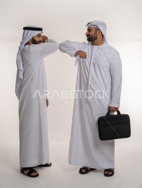 Following preventive measures against Corona virus, business agreement between businessmen, portrait of two Emirati Gulf Arab men wearing kandura, ghutra and mask and shaking hands with elbows, full body, white background