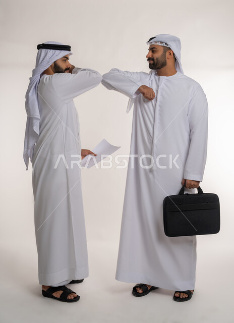 اتباع الاجراءات الوقائية ضد فايروس كورونا، اتفاق عمل بين رجال الاعمال، بورتريه لرجلان عربيان خليجيان اماراتيان يرتديان الكندورة والغترة و الكمامة ويتصافحان بالأكواع، الجسم بالطول الكامل، خلفية بيضاء