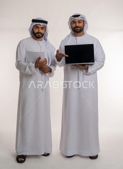 النظر الى الكاميرا بإيماءات السرور, مفهوم الادارة المتعاونة, اتباع الاجراءات الوقائية ضد فايروس كورونا, بورتريه لرجلين عربيين خليجيين اماراتيين يرتديان الغترة والكندورة والكمامة يحمل بيده الحاسب المحمول بشاشة سوداء ويشير اليه, الجسم بالطول الكامل, خلفية بيضاء