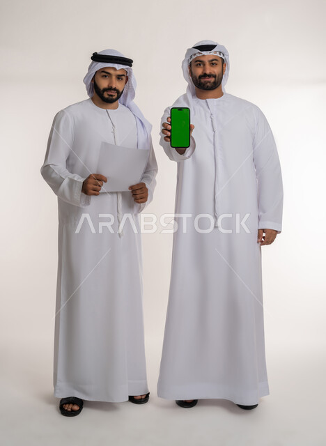 Looking at the camera with gestures of pleasure and admiration, using advanced technology and technology, making new partnership deals, portrait of two Emirati Gulf Arab employees wearing a ghutra, kandura and a mask, holding a mobile phone with a green screen, full-length body, white background