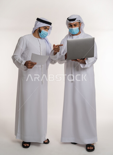مفهوم الإدارة المتعاونة، بورتريه لموظفين عربيين خليجيين اماراتيين يرتديان الغترة والكندورة والكمامة يتناقشان باستخدام الأوراق، اتباع الاجراءات الوقائية ضد كوفيد19, تحقيق أهداف الشركة وحل المشكلات و عقد صفقات شراكة جديدة، الجسم بالطول الكامل, خلفية بيضاء