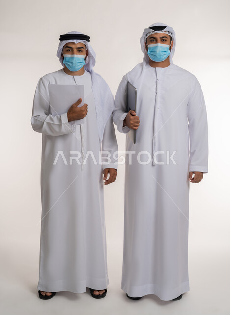 Making new partnership deals, achieving company goals and solving problems, collaborative management concept, portrait of two Emirati Gulf Arab employees wearing ghutra, kandura and mask looking at the camera with gestures of self-confidence and seriousness, following preventive measures against Covid-19, full body, white background