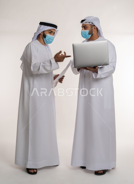 Achieving company goals, solving problems and making new partnership deals, collaborative management concept, portrait of two Emirati Gulf Arab employees wearing ghutra, kandura and mask discussing using papers, following preventive measures against Covid-19, full body, white background