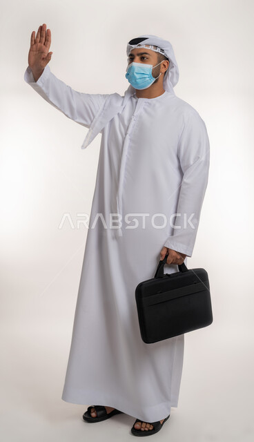 الحفاظ على المسافات وعدم الملامسة السطحية، بورتريه لرجل عربي خليجي اماراتي يرتدي الكندورة والغترة و الكمامة يحمل حقيبة العمل يرفع يده لتجنب المصافحة وعدم اقتراب الآخرين منه، اتباع الاجراءات الوقائية ضد فايروس كورونا، الجسم بالطول الكامل، خلفية بيضاء