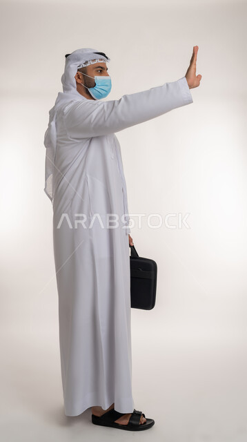 الحفاظ على المسافات وعدم الملامسة السطحية، بورتريه من الجانب لرجل عربي خليجي اماراتي يرتدي الكندورة والغترة و الكمامة يحمل حقيبة العمل يرفع يده لتجنب المصافحة وعدم اقتراب الآخرين منه، اتباع الاجراءات الوقائية ضد فايروس كورونا، الجسم بالطول الكامل، خلفية بيضاء