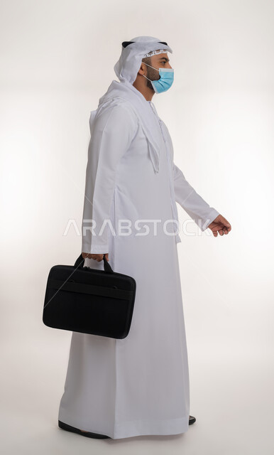 ايماءات تدل على الحركة والمشي, بورتريه لرجل أعمال عربي خليجي اماراتي يرتدي الكندورة والغترة و الكمامة الواقية ويحمل حقيبة العمل، الالتزام باجراءات الوقاية من فايروس كورونا، صورة الجسم بالطول الكامل، خلفية بيضاء