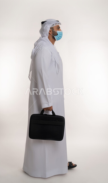 ايماءات تدل على الحركة والمشي, بورتريه لرجل أعمال عربي خليجي اماراتي يرتدي الكندورة والغترة و الكمامة الواقية ويحمل حقيبة العمل، الالتزام باجراءات الوقاية من فايروس كورونا، صورة الجسم بالطول الكامل، خلفية بيضاء