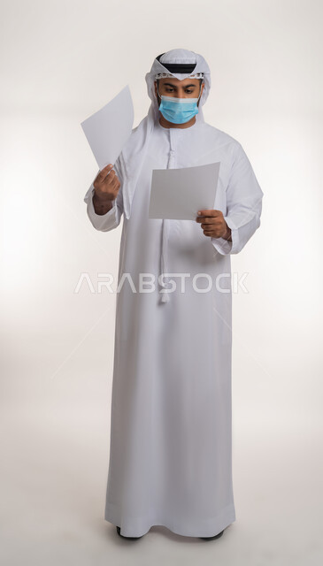 Stay safe from Covid-19, awareness panels to avoid the spread of infection, following precautionary measures to prevent the Corona virus, portrait of an Arab Gulf Emirati man wearing a ghutra and a kandura, wearing a mask, holding a white rectangular mockup in his hand, full-length body, white background