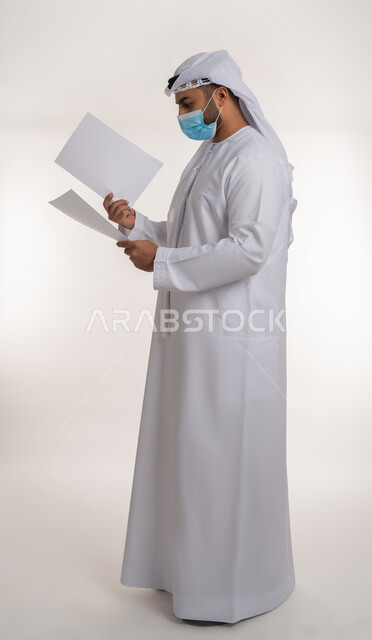 الحفاظ على السلامة من كوفيد 19، لوحات توعوية لتجنب إنتشار العدوى، اتباع الاجراءات الاحترازية للوقاية من فايروس كورونا، بورتريه من الجانب لرجل عربي خليجي إماراتي يرتدي الغترة والكندورة يضع الكمامة يمسك بيده موك اب مستطيل أبيض فارغ, الجسم بالطول الكامل، خلفية بيضاء