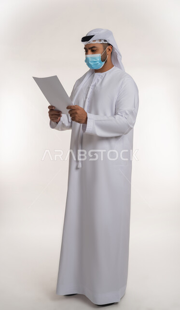 لوحات توعوية لتجنب إنتشار العدوى، الحفاظ على السلامة من كوفيد 19، اتباع الاجراءات الاحترازية للوقاية من فايروس كورونا، بورتريه لرجل عربي خليجي إماراتي يرتدي الغترة والكندورة يضع الكمامة يمسك بيده موك اب مستطيل أبيض فارغ, الجسم بالطول الكامل، خلفية بيضاء