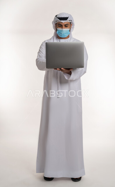 Spreading health awareness through the use of modern technical devices, working remotely to maintain safety, taking measures to prevent Covid-19, portrait of an Arab Gulf Emirati man wearing traditional dress and mask working on laptop, full body, white background