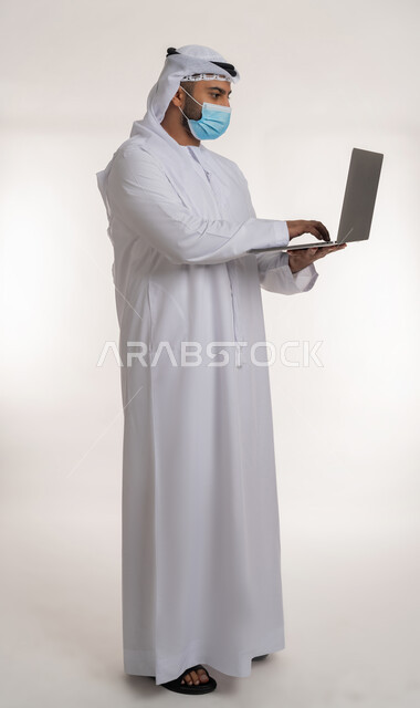 العمل عن بعد للحفاظ على السلامة,  نشر الوعي الصحي من خلال استخدام الأجهزة التقنية الحديثة, القيام بالإجراءات للوقاية من كوفيد 19, بورتريه من الجانب لرجل عربي خليجي اماراتي يرتدي الزي التقليدي والكمامة يعمل على الحاسب المحمول, الجسم بالطول الكامل, خلفية بيضاء