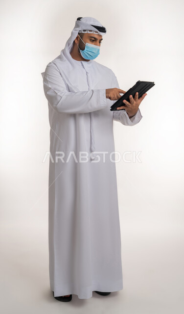 نشر الوعي الصحي من خلال استخدام الأجهزة التقنية الحديثة, القيام بالإجراءات للوقاية من كوفيد 19, مفهوم العمل عن بعد للحفاظ على السلامة, بورتريه لرجل عربي خليجي اماراتي يرتدي الزي التقليدي والكمامة يعمل على الجهاز اللوحي, الجسم بالطول الكامل, خلفية بيضاء