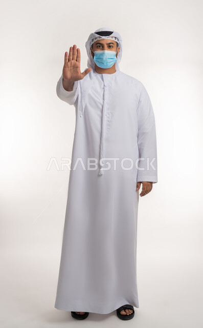 الالتزام بإجراءات الوقاية، تجنب انتشار العدوى بكوفيد 19، بورتريه لرجل عربي خليجي اماراتي يرتدي الكندورة والغترة والكمامة الواقية ينظر الى الكاميرا يرفع يده للاعلى بإيماءات التوقف وعدم الاقتراب، الجسم بالطول الكامل, خلفية بيضاء