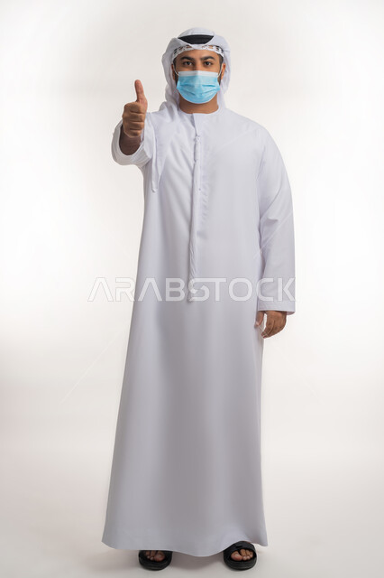 تجنب انتشار العدوى بكوفيد 19، النظر الى الكاميرا بتعابير الحذر، بورتريه لرجل عربي خليجي اماراتي يرتدي الكندورة والغترة والكمامة الواقية يرفع اصبع الابهام للاعلى بإيماءات القبول والرضا، الالتزام بإجراءات الوقاية، الجسم بالطول الكامل, خلفية بيضاء