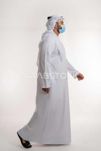 القيام بالإجراءات الاحترازية للوقاية من كوفيد 19, بورتريه من الجانب لرجل عربي خليجي اماراتي يرتدي الكندوره والغتره البيضاء والكمامة الوقائية بايماءات تدل على الحركة والمشي، الجسم بالطول الكامل، خلفية بيضاء