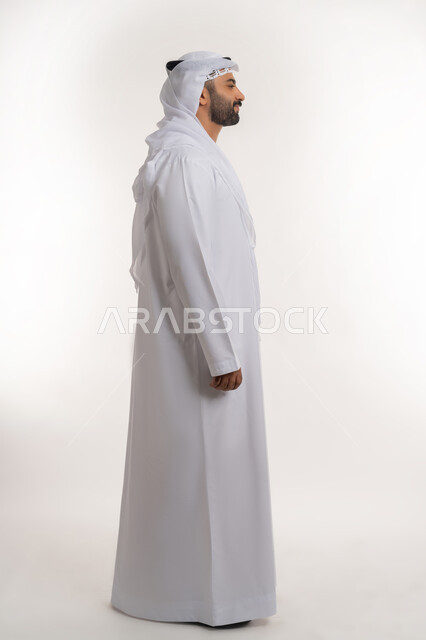 تعابير الثقه بالنفس و الجدية والحزم، مفهوم الاناقه والاهتمام بالمظهر الخارجي، بورتريه من الجانب لرجل عربي خليجي اماراتي يرتدي الكندوره والغتره البيضاء يقف بايماءات الرجولة، الجسم بالطول الكامل، خلفية بيضاء