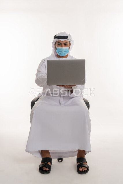 استخدام الأجهزة التقنية الحديثة لنشر الوعي الصحي والحد من انتشار الأمراض, القيام بالإجراءات الاحترازية للوقاية من كوفيد 19, مفهوم العمل عن بعد للحفاظ على السلامة, بورتريه لرجل عربي خليجي اماراتي يرتدي الزي التقليدي يضع الكمامة يعمل على الحاسب المحمول, خلفية بيضاء